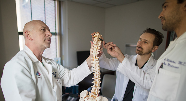 Three doctors hold and point to a model spine in an office setting