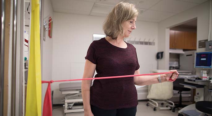 In an exam room, a woman pulls horizontally on pink exercise band